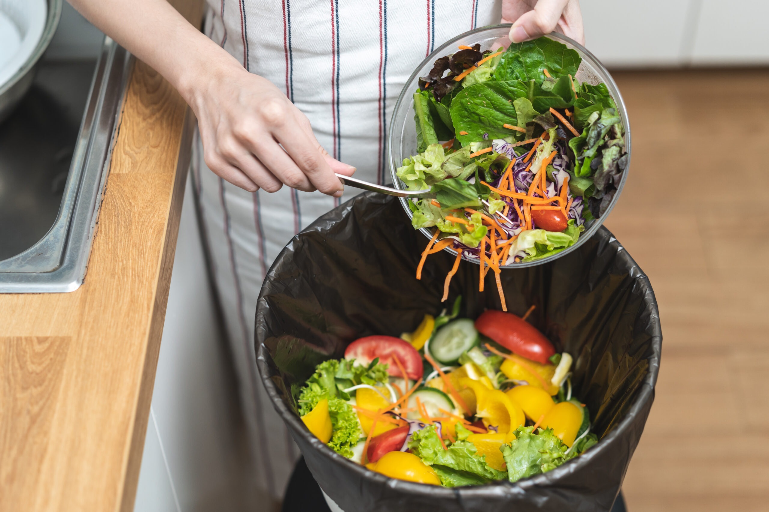 Une restauratrice vide un bol de salade fraîche remplie de légumes colorés dans une poubelle de cuisine, illustrant le gaspillage alimentaire domestique.